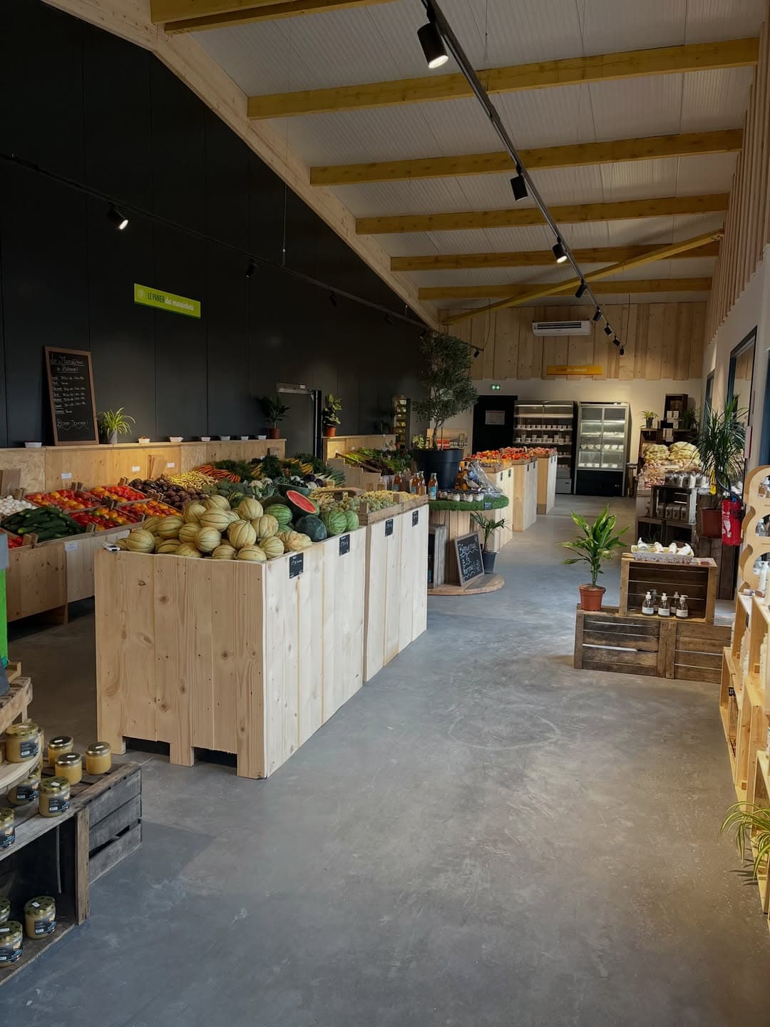 L'intérieur du magasin : étals en bois garnis de melons, pastèques et légumes, sous une charpente en bois apparente